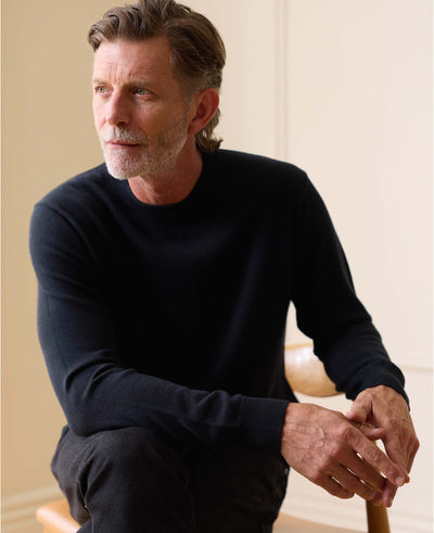 A man with graying hair and a beard wearing a dark navy long-sleeve knit sweater, sitting in a chair against a warm beige background.