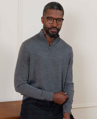 A man wearing a dark gray quarter-zip long-sleeve sweater and black glasses, standing against a white wall.