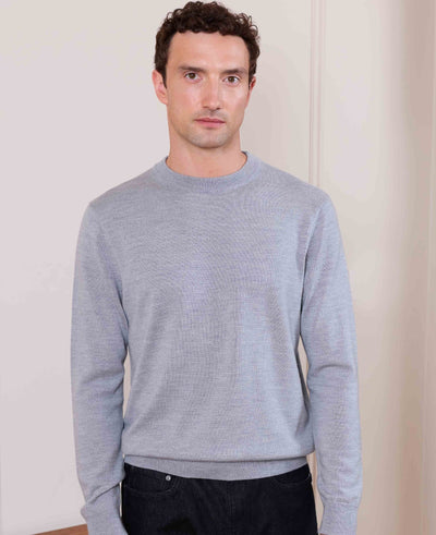 A man wearing a light gray ribbed knit crewneck sweater and dark pants, standing against a plain off-white wall.