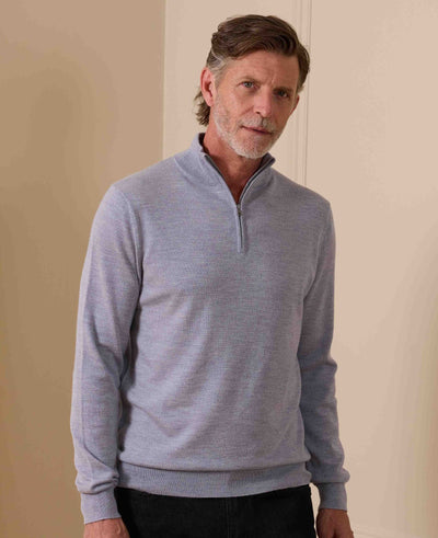 A man wearing a light gray quarter-zip knit sweater, standing against a neutral beige wall.