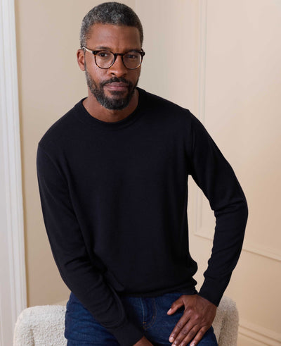 A man with short gray hair and a beard wearing black-rimmed glasses, a black long-sleeve crewneck sweater, and blue jeans, sitting in a soft white armchair against a beige paneled wall.