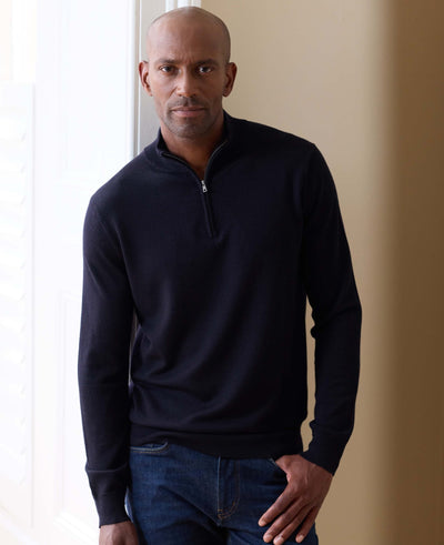 A man wearing a dark navy blue quarter-zip long-sleeve knit sweater and blue jeans, standing in a brightly lit indoor setting with a beige wall.